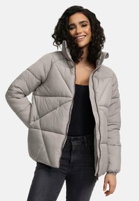 Lichtgrijze gewatteerde jas met een hoge kraag, voorzien van geometrische stiksels en ritsluiting. Draag het over een zwart topje, in combinatie met donkere jeans.