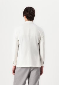 White textured blazer with a single vent at the back, long sleeves, and metal button accents. Worn over light gray pants.
