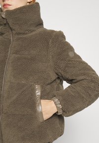 Hnědá fleecová bunda s vysokým límcem, zipovým zapínáním a lesklými světle hnědými doplňky na kapsách a manžetách. Texturovaný povrch a volnější střih.