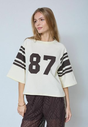 Jonge vrouw met lang blond haar, gekleed in een losse witte blouse met het nummer 87 en zwarte gestreepte mouwen, gecombineerd met donkere broek met patroon.