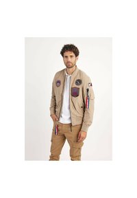 Veste bomber beige avec divers écussons, poignets côtelés et fermeture éclair. Associée à un pantalon cargo marron avec des poches latérales et des zips.