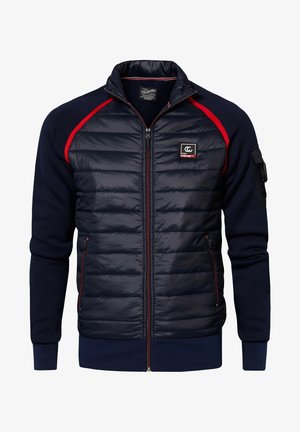 Niño con chaqueta acolchada azul marino con detalles en rojo, sudadera roja, vaqueros negros y zapatillas blanco y negro, de pie contra una pared lisa.