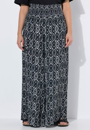 Kobieta w czarno-białych szerokich spodniach z wzorem diamentu, stojąca na białym tle, widoczna od talii do stóp.