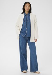 Žena v bielom pletenom kardigáne cez zapínanú denimovú košeľu a široké denimové nohavice, stojí s jednou rukou v vrecku na bielom pozadí.