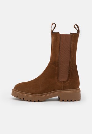 Brune ruskind ankelstøvle med elastiske sidepaneler og en pull tab, med en tekstureret gummisål og syninger langs kanten.