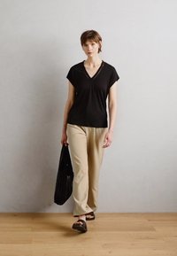 Top nero a maniche corte con scollo a V, pantaloni beige e sandali neri. Porta una borsa nera e si trova su un pavimento di legno contro una parete chiara.
