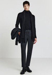 Dolcevita nero e sciarpa, pantaloni tailored scuri con una sottile texture, mentre tiene un giacca coordinata, abbinata a scarpe nere stringate.