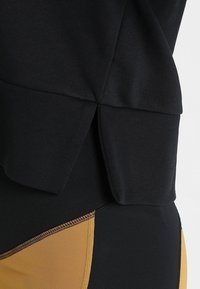Zwart sporttop van een gestructureerde stof, met een licht uitlopende zoom en een zijsplit. Gecombineerd met aansluitende zwarte en gele leggings.