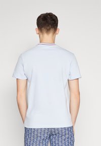 Světle modré polo tričko s krátkým rukávem, které má červený límeček. Tkanina vypadá hladce a je koncipována ve fitted stylu, z pohledu zezadu.