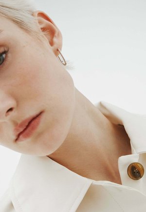 Primer plano de una persona con cabello corto rubio que lleva un arete en forma de alfiler de seguridad plateado y una camisa de color crema con un botón marrón.