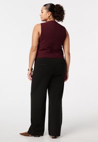 Femme avec une queue de cheval frisée portant un haut sans manches bordeaux, un pantalon large noir, des chaussures à talons pointus en imprimé léopard et des bijoux en or, se tenant de dos.