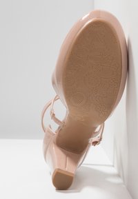 Chaussure à talon haut rose nude brillant avec bride à la cheville, montrant la semelle inférieure embossée de motifs floraux.