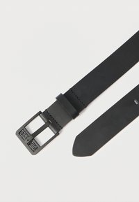 Čierny kožený opasok s matným povrchom a štvorcovou čiernou kovovou prackou. Má čistý dizajn s kontrastným prešívaním pozdĺž okrajov.