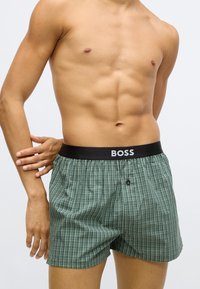 Zöld kockás boxeralsó fekete gumiszalaggal, melyen a "BOSS" logó látható. Könnyű anyagból készült, elülső gombos részlettel.