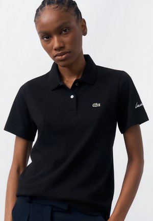 Vrouw met gevlochten haar die een zwarte Lacoste polo draagt met het groene krokodillenlogo van het merk en de tekst "Lacoste" op de mouw.