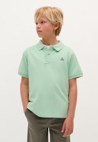 Maglietta polo verde chiaro con colletto, maniche corte e taschino frontale. Presenta un piccolo logo a forma di teschio di colore più scuro sul petto.