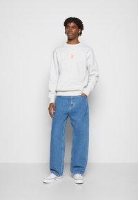 Siva sweatshirt z oranžnim logotipom, sproščen kroj, v kombinaciji s širokimi modrimi kavbojkami. Bele superge dopolnjujejo outfit.
