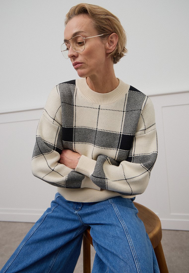 Beige sweater met zwart-wit ruitpatroon, met ribgebreide manchetten en halslijn. Losse pasvorm, gecombineerd met blauwe wijde jeans.