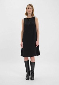 Robe noire sans manches avec une coupe droite, en tissu lisse, et une longueur jusqu'aux genoux ; associée à des bottes noires jusqu'au genou et à des bijoux en or simples.