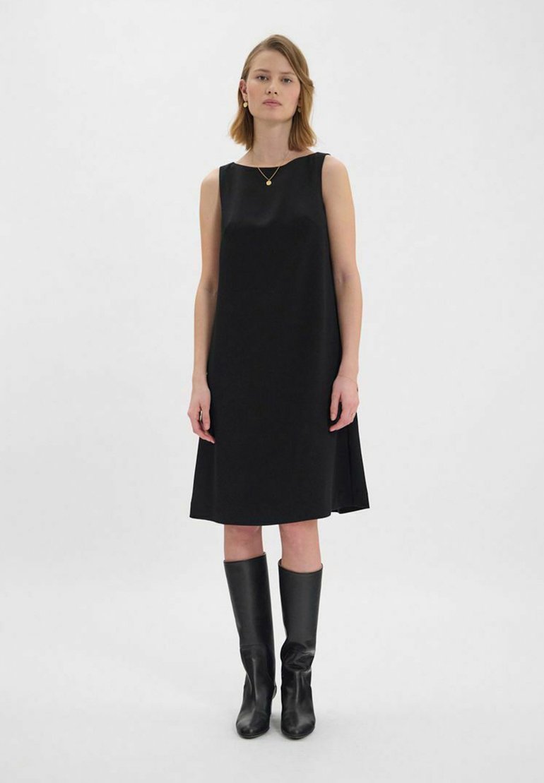 Robe noire sans manches avec une coupe droite, en tissu lisse, et une longueur jusqu'aux genoux ; associée à des bottes noires jusqu'au genou et à des bijoux en or simples.