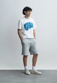 Λευκό μπλουζάκι με μπλε γραφικό κείμενο, συνδυασμένο με γκρι σορτς από φλις. Το μοντέλο φορά λευκά αθλητικά παπούτσια και λευκές κάλτσες μήκους μέχρι τον αστράγαλο πάνω σε γκρι δάπεδο.