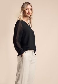 Blusa nera a scollo a V con maniche lunghe trasparenti, abbinata a pantaloni a gamba larga beige chiaro, che mostrano una silhouette rilassata e una morbida texture.