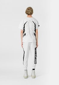 Camiseta de algodón blanca con gráficos de alas negras en la parte posterior, combinada con pantalones deportivos blancos que tienen acentos y etiquetas negras en los laterales.