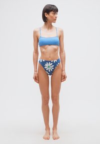 Γυναίκα ελάχιστα ντυμένη με μπλε μπικίνι που έχει μοτίβο κυμάτων στο επάνω μέρος και φλοράλ σχέδιο σε ψηλόμεσο κάτω μέρος, κοιτάζει προς τα δεξιά της.