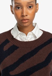 Brauner Strickpullover mit dunkelblauen abstrakten Streifen, mit geripptem Kragen, über einem weißen Hemd mit dünnen vertikalen Streifen getragen.