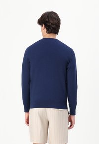 Tummansininen neulepaita, jossa on pyöreä kaula-aukko, pitkät hihat ja ribattu hihansuu, yhdistettynä beigekirjaviivaisiin shortseihin. Sujuva pinta.