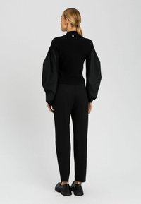 Pullover nero in maglia con maniche in tessuto gonfie e un piccolo bottone accento sulla parte posteriore, abbinato a pantaloni neri su misura e scarpe nere chunky.