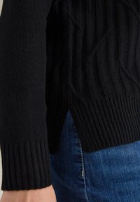 Pull en tricot noir avec un motif texturé, des poignets côtelés et des fentes sur les côtés, associé à un jean en denim bleu.