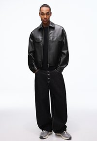 Junger Mann, der eine schwarze Lederjacke, lockere schwarze Jeans mit Knopfleiste und graue Turnschuhe trägt, steht mit den Händen in den Taschen vor weißem Hintergrund.