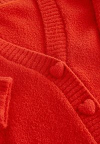 Rdeč pulover iz mehke, puhasto materiala. Ima rebrast ovratnik in gumbe v obliki srca. Teksturirana površina kaže topel, živahen odtenek.