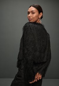 Chaqueta negra texturizada hecha de material peludo y con flecos, con toques brillantes y un ajuste relajado. Llevada sobre pantalones negros.