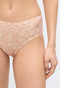 Γυναίκα που φοράει ελαφρύ ροζ εσώρουχο με μοτίβο λεοπάρ, που φαίνεται η μέση, το δεξί χέρι και το πάνω μέρος του μηρού της, πάνω σε λευκό φόντο.