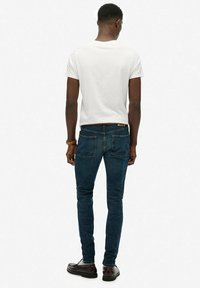 Jeans ajustados de color azul oscuro con una textura suave, combinados con una camiseta blanca de manga corta y zapatos marrones. Vista trasera del conjunto.