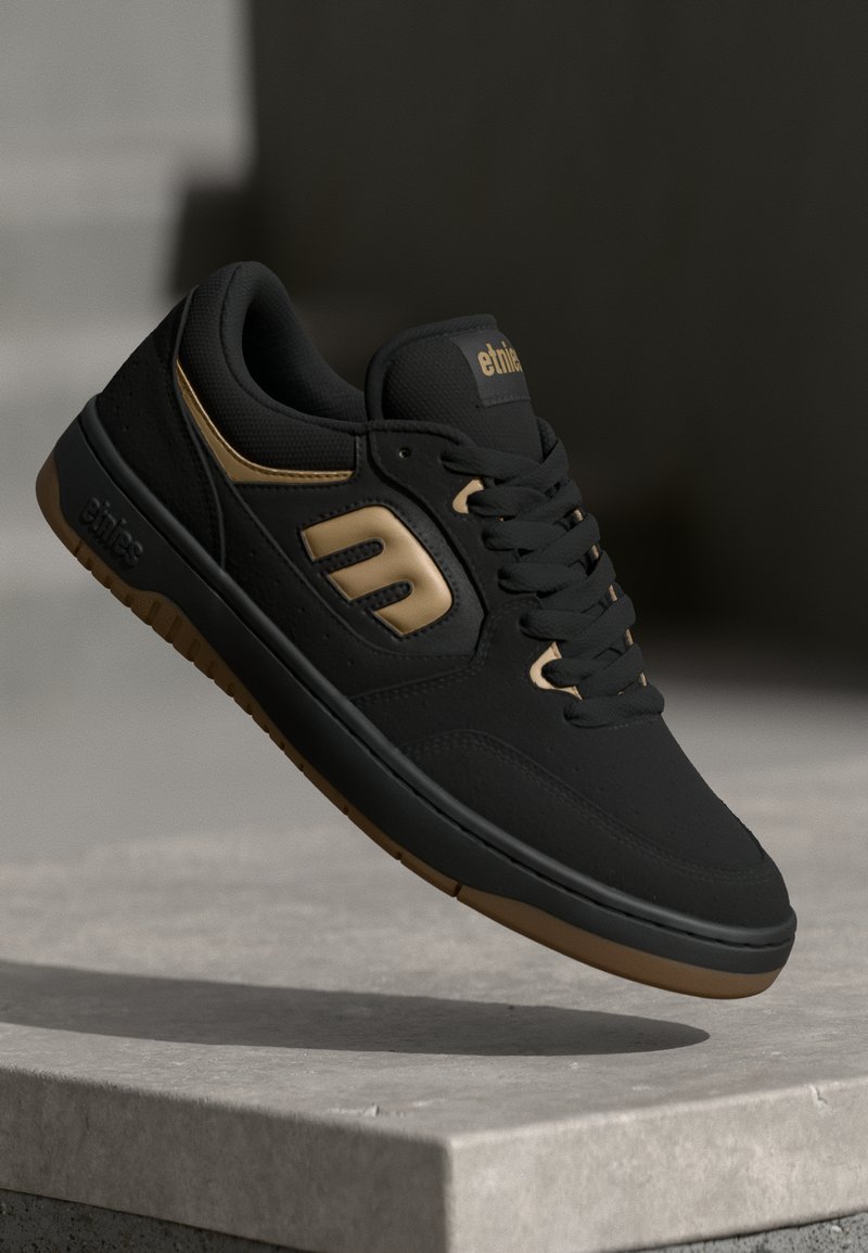 Zapatilla negra con acentos dorados, parte superior de cuero suave y malla, suela de goma, punta redonda y logotipo prominente en el lateral.