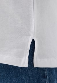 White cotton shirt hem with side slit detail, revealing blue denim fabric beneath. Smooth texture, clean stitching along the edges.