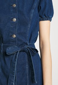 Blå denimklänning med puffiga korta ärmar, metallknappar framtill, och knytbart midjebälte på en ljushyad arm visad från axel till höft.