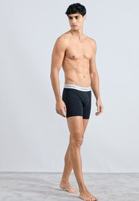 Fekete mintás boxeralsó, fehér logós derékszíjjal, testhez simuló kialakítással és sima anyaggal, férfi modell viselésében, semleges háttér előtt.
