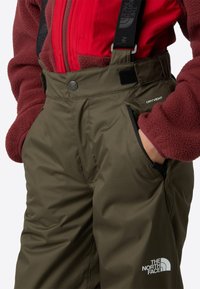 Olive green waterproof pants with adjustable black suspenders and side pockets. Features branding logo at the lower leg and smooth texture.