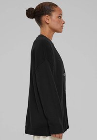 Svart oversized cardigan med ribbetkant og mansjetter, rund hals og knappdetaljer. Laget av mykt strikkemateriale.