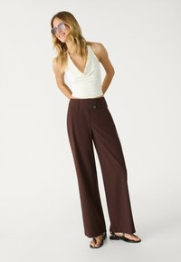 Bruna vida byxor med hög midja och tre knappljus, kombinerade med en vit halterneck-topp och svarta platta sandaler.