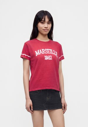 Nuori nainen, jolla on pitkät tummat hiukset ja joka on pukeutunut punaiseen "Marseille 1982" t-paitaan ja tummaan farkkuhameeseen, seisoo yksiväristä valkoista taustaa vasten.