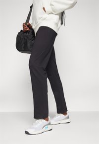 Pantalones deportivos negros con diseño de pierna recta, combinados con zapatillas blancas y grises con detalles en azul y una bolsa texturada en la mano.