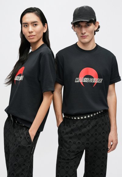 T-shirts de algodão pretas apresentam um logótipo de crescente vermelho e o texto "MARINE SERRE". Combinadas com calças pretas com padrão e um cinto com tachas.