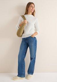 Donna in maglione bianco, jeans blu e sneakers sta in piedi tenendo una borsa a spalla imbottita verde oliva, sorridendo e guardando a destra.