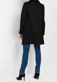 Černý vlněný kabát s přizpůsobeným střihem, klopovým límcem a bočními kapsami, doplněný o modré džíny a černé kotníkové boty na podpatku.