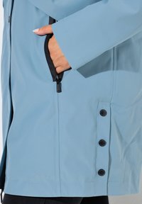 Chaqueta azul celeste con textura suave, bolsillo con cremallera negra y botones metálicos en el dobladillo. Presenta un corte recto y mangas largas.